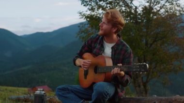 Romantic millennial sing nature in mountains. Close up smiling guy relax on travel camp. Young guy practice music play acoustic guitar outdoors. Relaxed tourist chill on green hill. Leisure concept.