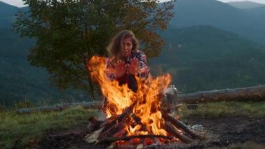 Beautiful woman warm hands on campfire. Wanderlust traveler enjoy mountains evening close up. Relaxed tourist sit log in forest. Traveling hipster on social distance tourism activity. Leisure concept.