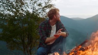 Smiling traveler warm fire on camp. Joyful millennial rest in mountains forest closeup. Happy guy warming hands by bonfire outdoors. Redhead tourist enjoy peaceful evening. Healthy lifestyle concept.