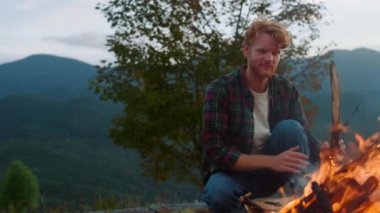 Active traveler camp forest in mountains. Close up redhead hiker sit bonfire on nature evening sunset. Cheerful camper throw log wood in fire. Young tourist spend weekend. Leisure relax travel concept