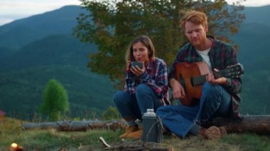 Relaxed lovers play guitar on nature view. Hikers couple spend vacation outdoors close up. Young family enjoy camping on green hill mountains. Happy tourists play guitar by campfire. Leisure concept.