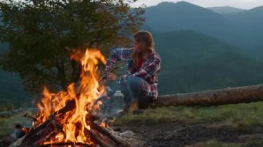 Active woman spend evening by bonfire. Young hiker play acoustic guitar in mountains green hill close up. Positive millennial rest on nature landscape. Traveler musician perform outside. Relax concept