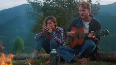 Wanderlust couple spend weekend in mountains. Young family enjoy camp evening outdoors closeup. Thoughtful travelers play music instrument guitar by bonfire. Tourism youth lifestyle leisure concept.