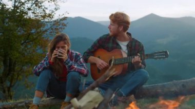 Young tourists enjoy campfire in mountains. Close up hipsters couple play music on nature hill. Joyful campers drink tea on summer evening. Relaxed travelers spend holiday together. Outdoors concept.