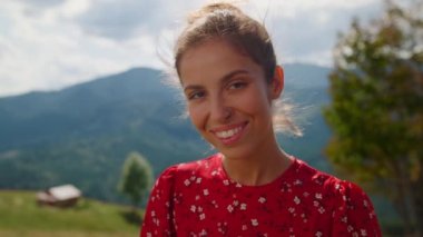 Closeup smiling pretty woman posing in front mountains summer holiday. Portrait of happy girl looking camera standing green meadow. Positive lady wearing red dress relax outdoors on sunny day.