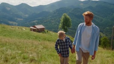 Happy man with son walking on mountain slope sunny day. Red hair father with boy holding hands on family trip outdoor. Young parent with child coming up green hill on summer weekend. Vacation concept.