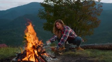 Thoughtful hipster travel camping spend vacation in mountains campfire nature. Close up young woman throw wood in bonfire outside. Active tourist enjoy fire in evening. Adventure lifestyle concept.