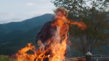 Camping hipster watch fire in nature forest. Happy guy sit bonfire in mountains close up. Wanderlust tourist traveling on beautiful landscape. Positive traveler enjoy evening. Activity leisure concept