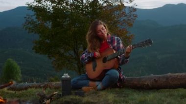 Closeup musical traveler perform song play acoustic guitar in mountains nature. Smiling girl relax on wanderlust activity journey. Attractive hipster enjoy campfire at evening. Leisure music concept.