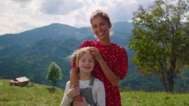 Smiling woman stroking girl head standing in front mountains sunny day close up. Happy mother with daughter cuddling together on green meadow. Joyful mom with cute child enjoying summer family holiday