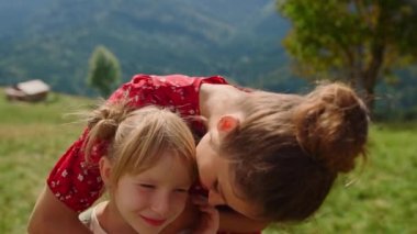 Close up loving mother kissing daughter on green meadow sunny day. Portrait of happy woman hugging with cute blonde child outdoor. Smiling mom spending summer vacation with girl in mountains.