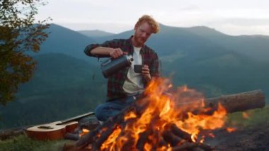 Relaxed tourist enjoy camping lifestyle by bonfire. Chill guy drink pour tea outside in mountains closeup. Young man hold thermos on hiking vacation. Redhead traveler spend evening. Adventure concept.