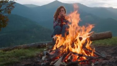 Closeup relaxed woman camping on nature. Beautiful tourist look bonfire in mountains trip. Happy girl drink tea by burning log bonfire. Carefree traveler rest on evening. Wanderlust concept leisure.