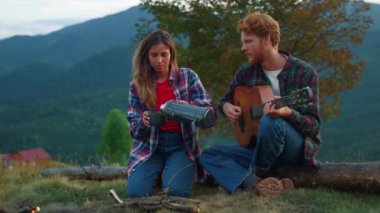 Family couple enjoy mountains on camping. Close up two travelers play guitar song outdoors on green hill. Young campers drink hot thermos tea on nature journey trip. Wanderlust leisure relax concept.