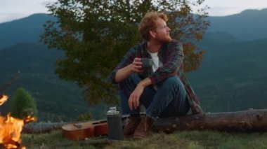 Joyful hiker relax bonfire in mountains green hill. Close up camper enjoy drink outside by evening campfire. Happy traveler sit log on summer weekend nature. Active holiday recreation leisure concept.
