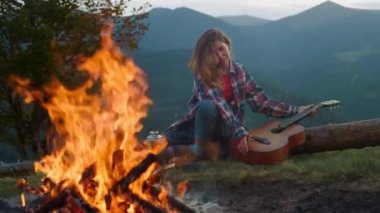 Attractive musician travel mountains. Young woman enjoy burning campfire closeup. Joyful girl drink hot tea at tourism camping site. Beautiful millennial hiker spend holiday outside. Trip concept.