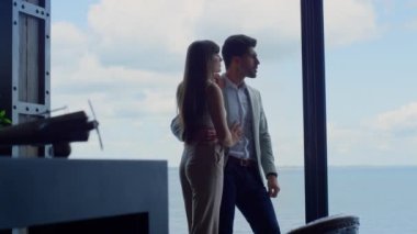 Sensual couple enjoying ocean view at hotel. Newlyweds talking spending vacation at panoramic window place. Handsome man hugging woman by waist. Lady and guy embracing together. Honeymoon concept