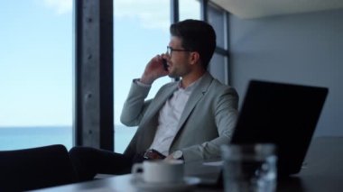 Positive professional talking smartphone at sea view workplace. Businessman having conversation on cellphone in luxury office. Joyful boss director call phone in panoramic window conference hall 