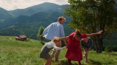 Happy family spreading hands like wings playing outdoor sunny day. Playful couple with children having fun running on green hill. Positive parents enjoy time together at summer vacation in mountains.