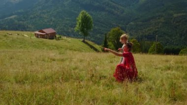 Carefree mother sitting near daughter on meadow sunny day. Happy woman relaxing with cute girl in mountains enjoying summer vacation. Young mom in red dress pointing finger at beautiful nature views.