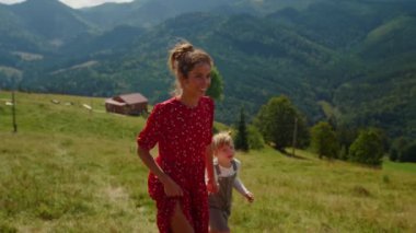 Happy woman showing daughter mountains views sunny day. Smiling mother holding girl hand walking on beautiful meadow. Cheerful mom relaxing with cute child on green hill. Family rest concept.