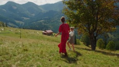 Back view of lovely mother with daughter going down green hill. Unknown woman with cute girl holding hands walking green grass in mountains. Young mom spending family holiday outdoors with child.