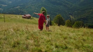 Back view of young woman walking with daughter on green hill. Unknown mother looking at mountains panorama with little girl outdoors. Lovely child with mom enjoying summer vacation in mountains.