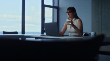 Focused businesswoman drinking water in empty office room. Ceo overworking alone creating corporate report surfing internet in glass office. Successful financial analyst look using laptop computer