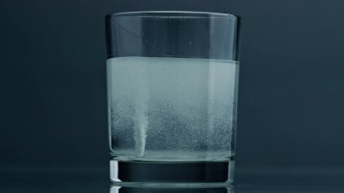 White pill dissolving glass at dark background closeup. Effervescent tablet flow in soda sparkling water transparent cup. Hangover disease treatment bubbling in glassware. Fizzy refreshing drink.