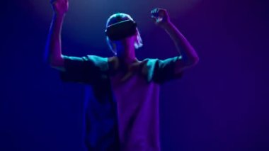 Millennial man partying with invisible 3D avatar. Virtual reality goggles person exciting playing neon background. VR-box youngster moving relaxing slow motion. Modern technological experience concept