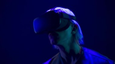 Focused VR headset man watching 3d movie in neon light closeup. Blonde gamer looking goggles over multicoloured background. Augmented reality helmet guy enjoying future technologies alone slow motion