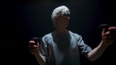 Teenager playing augmented reality game in dark room. Focused blonde man using console controllers in light. Guy explore immersing vr simulation in futuristic goggles. Interacting metaverse technology