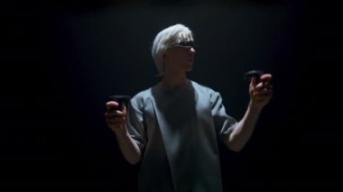 Cyber player enjoy augmented reality in futuristic headset. Guy use controllers experiencing vr simulation in dark room. Focused blonde man gesturing select objects in light. Gaming technology concept