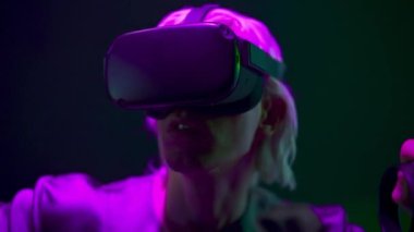 Amazed man playing videogame with remote controllers closeup. Focused gamer using joysticks and VR goggles at neon light place. Blonde headset guy interacting digital interface alone. Future concept 