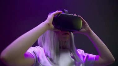 Milenyum kızı VR gözlüklerini yakın plan çekiyor. Etkilenmiş kadın yeni üç boyutlu simülasyon deneyimini yavaş çekime hazırlıyor. Kadın oyuncu eğlence sektörünün renkli neon siber punk arka planından zevk alıyor.