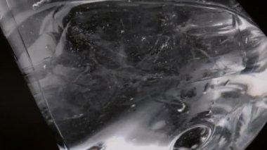 Saf suyla dolu şeffaf şarap kadehi ve buz küpleri. Donmuş bloklar, berrak sıvıyla dolu temiz bardakta yavaşça dönüyor. Siyah arka planda soğuk, ferahlatıcı bir içecek kadehi..