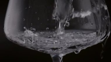 Siyah arka planda şeffaf kadehten temiz su sıçrıyor. Kristal içecekler şarap bardağına ağır çekimde dökülüyor. Fırtınalı sıvı yüzey cam içinde dönüyor. Organik içecek.