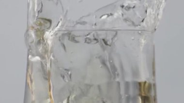 Şeffaf kadehe beyaz şarap damlatıyor. Kristal şarap kadehinin içine buz küpü düşüyor. Altın alkol damlacıkları beyaz arka plana dökülüyor..