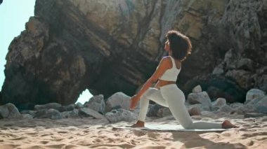 Kayalık Ursa plajı güneşli sabahında esnek bir kadın hafif hareketli poz veriyor. Genç yogi kızı, açık havada hasat eğitiminde hilal ay asanası yapıyor. Esmer kadın yoga savaşçısı pozu veriyor..
