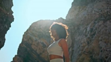 Yüksek kayalıklı Ursa plajı yakınlarında yoga yapan sportif bir kadının üzerinde parlayan yaz güneşi. Gülümseyen Afrikalı Amerikalı kız ellerini havaya kaldırarak uzanıyor. Rahatlamış sporcu kadın doğa üzerinde egzersiz yapıyor..