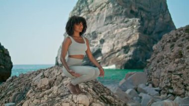 Sıska kız, elleri başının üstünde çömelmiş kayalık deniz kenarında yoga pozu veriyor. Afrika kökenli Amerikalı sporcu kadın Ursa sahilinde denge için Asana egzersizi yapıyor. Barış uyumu kavramı.
