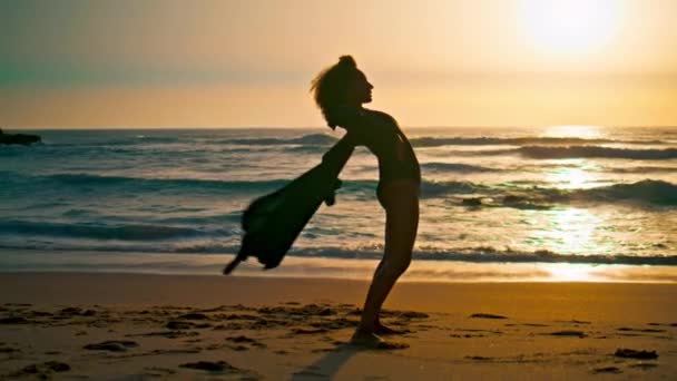日出日落时 轮廓柔韧的女人弯腰向后站在沙滩上 在自然夏日的早晨 放松了性感女孩伸展身体的运动 黑色的长袍神奇地在风中飘扬 — 图库视频影像