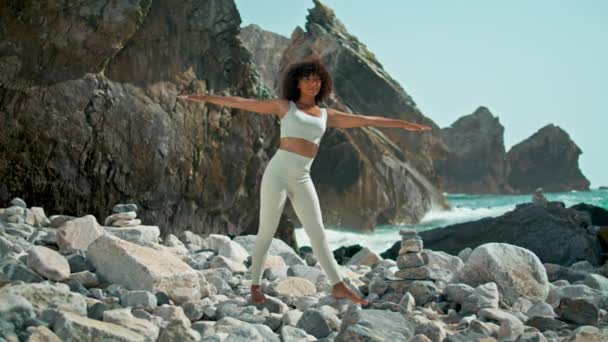 静下心来 在美丽的岩石上的乌尔沙海滩上练习胡萝卜素 做瑜伽的非洲裔美籍女孩在海浪前摆姿势 阳光灿烂 身材苗条的女运动员在户外伸展身体 — 图库视频影像