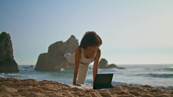 アフリカ系アメリカ人の女性は 砂のビーチの朝の時間にモダンなタブレットを使用してヨガのオンラインレッスンを見て 日の出には畳の上で立ちアサナを練習するスポーティ少女 自然の上で体を伸ばすスポーツ女性 — ストック動画