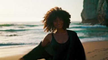 Güneşin doğuşunda kumsalda yürürken kamerada flört eden neşeli bir kadın. Mutlu Afro-Amerikalı kız deniz kıyısında eğlenceli bir yaz gezintisi yapıyor. Neşeli bayan sabah vakti dinleniyor..