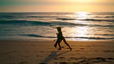 Sahilde gün doğumunda dans eden enerjik bir kadının silueti. Zarif kız ıslak deniz kıyısında baştan çıkarıcı bir şekilde hareket ediyor. Bayan dansçı el kaldırıyor. Kanat çırpan pareo ile büyüleyici bir performans sergiliyor..