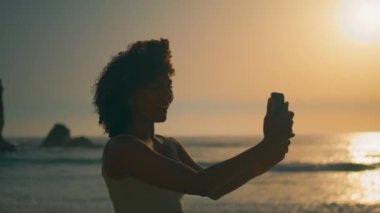 Güzel Afro-Amerikalı kız gün doğumunda kıyıda durup kameraya gülümsüyor. Çekici, mutlu bir kadın, doğal bir yaz akşamı selfie çekiyor. Tasasız kadın akıllı telefondan fotoğraf çekiyor..