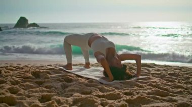 Güneşin doğuşunda kumsalda Urdhva Dhanurasana antrenmanı yapan sportif bir kadın. Afrika kökenli Amerikalı sporcu bir bacağı deniz kıyısında yüksekte poz veriyor. Esnek kız doğada yoga eğitimi alıyor..