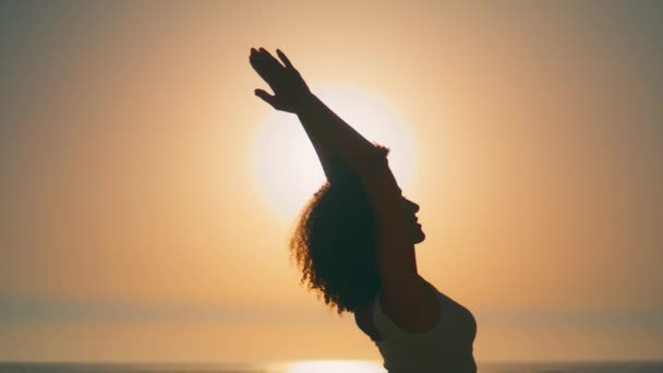若い女性のシルエット雲のない空に夏の朝のクローズアップ立って手を上げる 太陽の光の下で顔を置くリラックスした女の子は海岸で日の出を楽しんでいます 穏やかな女性ヨガの瞑想を練習 — ストック動画