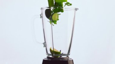 Dilimlenmiş vitamin sebzeleri çok yavaş çekimde blender kasesine düşer. Yeşil lezzetli sebzeler, beyaz arka planda cam karıştırma kabını dolduran meyve otları. Elmalı lahana kereviz salatalıklı maydanoz karışımı.